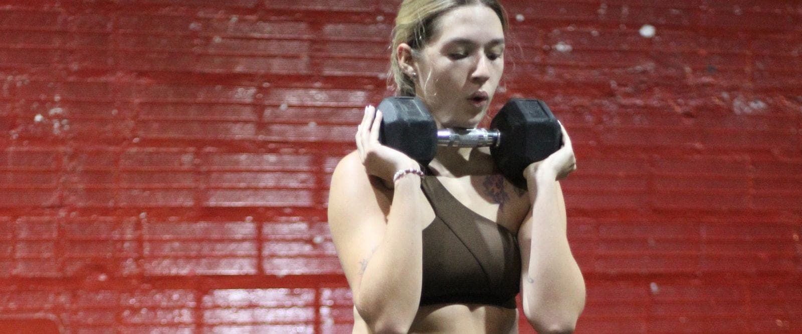Una atleta sostiene una mancuerna a la altura del pecho frente a la pared roja de BullBox.